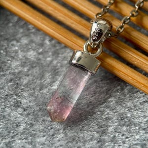 May include: A pendant necklace featuring a translucent, pale pink, hexagonal crystal point suspended from a silver-toned metal bail. The bail is stamped with the text 925, indicating sterling silver, and is attached to a delicate silver-toned chain.