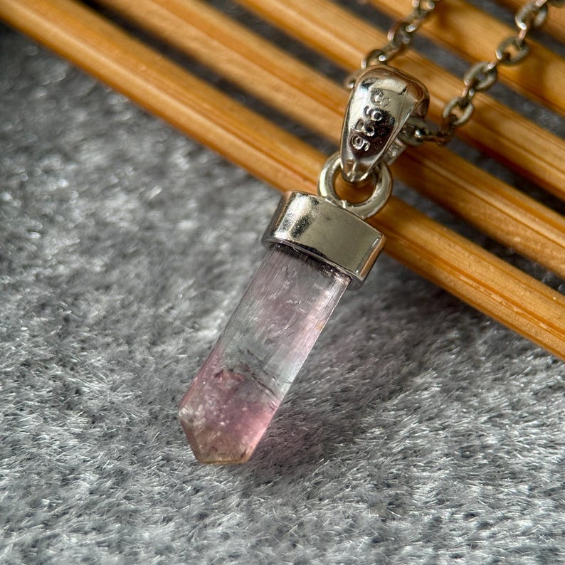May include: A pendant necklace featuring a translucent, pale pink, hexagonal crystal point suspended from a silver-toned metal bail. The bail is stamped with the text 925, indicating sterling silver, and is attached to a delicate silver-toned chain.