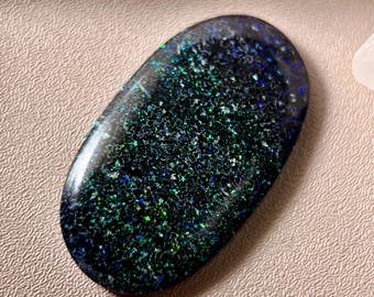 May include: An oval-shaped, polished gemstone with a dark base color. The stone is speckled with vibrant green, blue, and silver flecks, creating a shimmering, iridescent effect. The surface is smooth and reflective, showcasing the natural patterns.