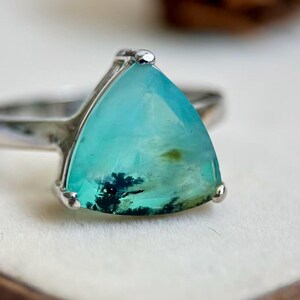 May include: A silver ring featuring a large, triangular, faceted teal gemstone. The stone displays internal moss-like black dendrite inclusions and a translucent quality. The metal band is polished silver, holding the stone with three prongs.