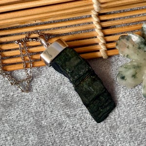 Könnte beinhalten: Ein dunkelgrüner, rechteckiger, texturierter Rohstein-Anhänger an einer dünnen silberfarbenen Metallkette. Der Stein hat eine raue, geschichtete Oberfläche und ist mit einer polierten silberfarbenen Metallkappe oben befestigt.