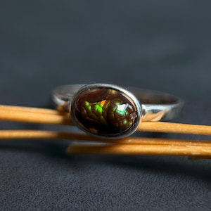 May include: A silver ring featuring an oval-shaped fire agate gemstone. The stone displays a dark brown base with iridescent flashes of green and orange light. The metal band is smooth and polished, encircling the cabochon-cut gemstone securely.