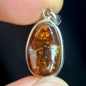 May include: A teardrop-shaped fire agate pendant set in a polished silver bezel. The gemstone features a translucent, deep reddish-brown base with iridescent, golden-orange botryoidal inclusions that create a textured, bubbly appearance.
