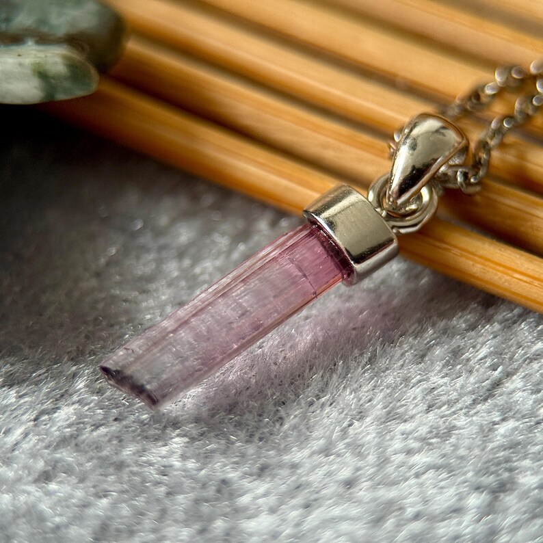 May include: A pendant necklace featuring a long, slender, translucent pink tourmaline crystal point held by a polished silver-toned metal bail. The crystal has a vertical striated texture and a rectangular shape, suspended from a matching silver-toned chain.