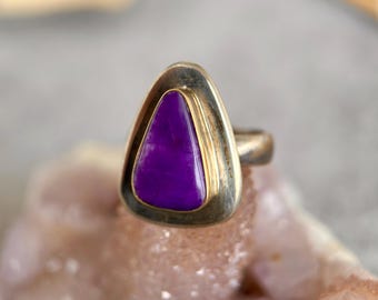 May include: A ring featuring a triangular, vivid purple stone set within a gold-toned bezel. The ring has a silver-coloured band. The ring is displayed on a light pink surface.