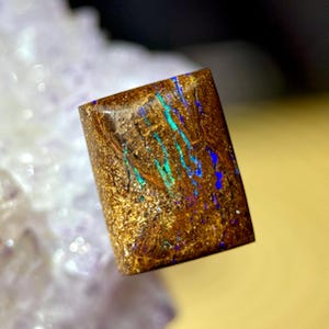Peut inclure: Une pierre d'opale rectangulaire et polie, avec une couleur de base brun terreux riche. La pierre présente des paillettes dorées scintillantes et des stries vibrantes de bleu et de vert-bleu, créant un effet visuel saisissant. L'opale est une pièce naturelle et unique.
