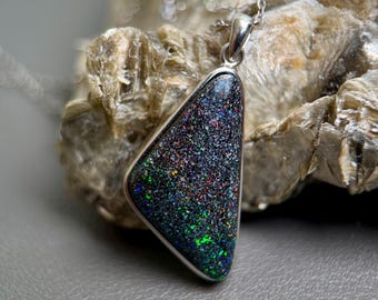 May include: A triangular pendant featuring a dark, textured stone with iridescent flecks of green, blue, and red. The stone is set in a silver-toned metal frame and hangs from a delicate silver-toned chain, showcasing a vibrant play of color.