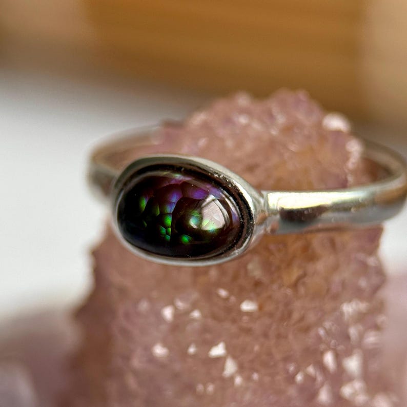 May include: A silver ring with an oval gemstone. The gemstone has a dark, iridescent quality, with green, purple, and black hues. The ring is displayed on a light pink, textured surface. The ring is a piece of jewellery.