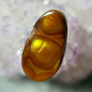 May include: A polished, oval-shaped gemstone with a warm, amber color and internal patterns. The stone has a glossy surface and displays concentric circles and a bright, yellow-green area, creating a unique, eye-catching appearance. The gemstone is likely used for jewelry.