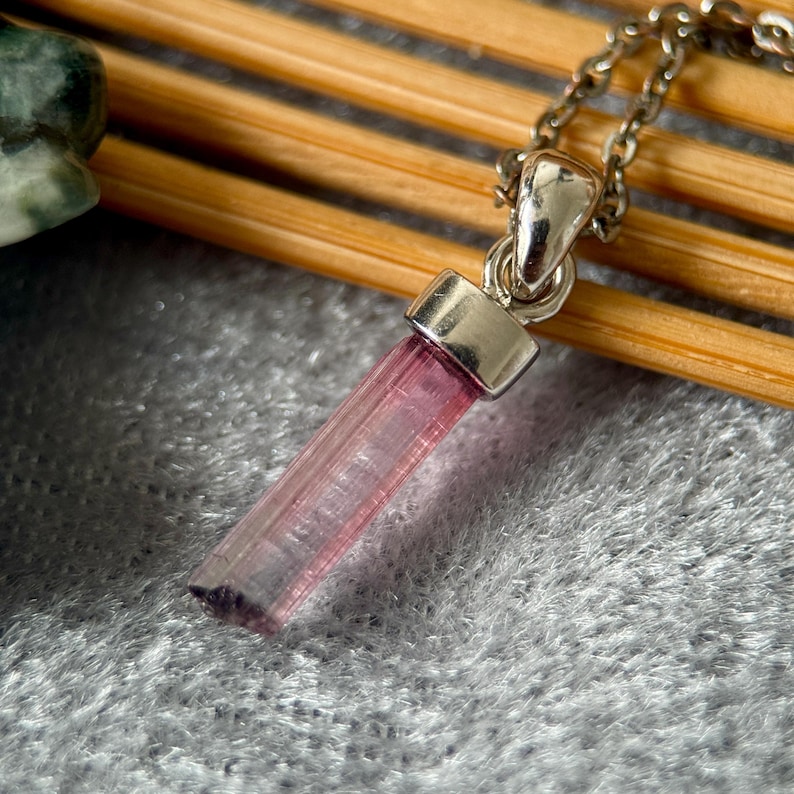 May include: A pendant necklace featuring a raw, cylindrical pink tourmaline crystal suspended from a silver-toned metal bail and a matching link chain. The translucent gemstone displays a gradient of light pink to a deeper, darker purple-toned base.