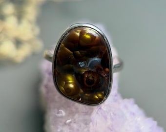 May include: A ring featuring a polished, irregular, oval-shaped fire agate gemstone set in a smooth silver-toned metal band. The stone displays deep brown, amber, and golden iridescent orb-like inclusions throughout its translucent surface.