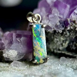 May include: A rectangular pendant featuring a dark, iridescent opal stone with vibrant flashes of green, blue, and yellow light. The stone is set in a silver-toned metal frame with four prongs at the corners and a circular bail at the top.