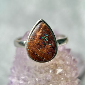 May include: A silver ring featuring a teardrop-shaped boulder opal gemstone. The stone has a dark brown, textured surface with vibrant, iridescent flashes of blue and green light embedded within the natural cracks and patterns of the gem.
