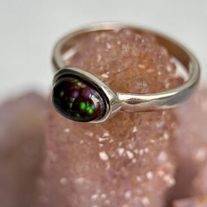 May include: A silver ring featuring a teardrop-shaped gemstone with a mix of colours, including green, purple, and brown. The ring is set on a pinkish-orange surface. The silver band contrasts with the gemstone's colours.