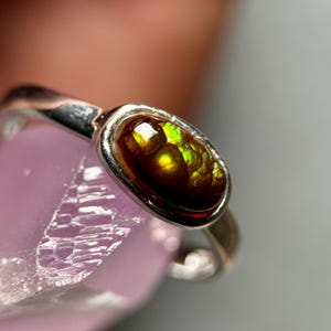 Op de afbeelding: Zilveren ring met een ovale edelsteen met bruine, groene en gele tinten. De steen is gezet in een bezel setting. De ring wordt getoond op een lichtpaars oppervlak.