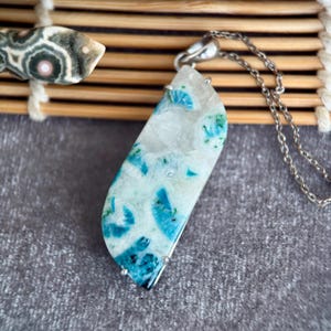 Puede incluir: Un collar con un colgante de piedra rectangular, blanco con patrones azules y verdes, suspendido de una cadena plateada. El collar se exhibe sobre una superficie gris.