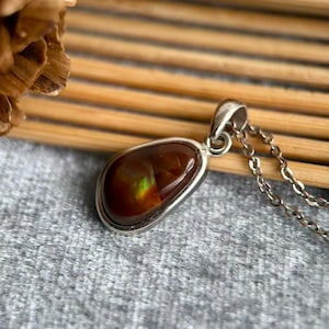 Op de afbeelding: Een ketting met een druppelvormige hanger met een gepolijste, roodbruine edelsteen. De steen heeft een subtiele, iriserende glans van groen en geel. De hanger is gezet in een zilverkleurig metalen frame en hangt aan een fijne ketting.