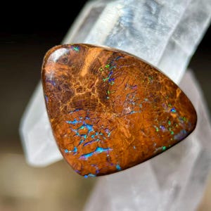 Peut inclure: Une pierre d'opale triangulaire polie, de couleur de base marron riche. La pierre présente des motifs et des veines complexes avec des éclairs d'irisation bleue, verte et jaune. L'opale est posée sur un fond cristallin clair.