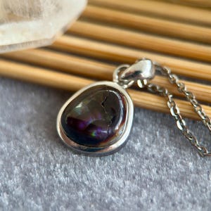 Op de afbeelding: Een zilveren hangerketting met een druppelvormige, iriserende zwarte edelsteen. De edelsteen heeft een gebarsten uiterlijk en is gezet in een zilveren bezel. De ketting is ook zilver.