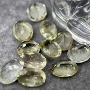 May include: A selection of oval-cut gemstones in pale yellow-green hues. The gems are scattered on a grey surface, with a clear glass object partially visible. The stones have a polished, reflective surface. The gems are likely to be measured in millimetres.