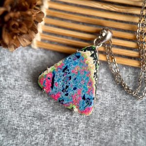 May include: A colorful pendant necklace with a triangular, abstract design. The pendant features a mix of blue, pink, and green hues with black accents. The necklace has a silver chain and clasp. The pendant is approximately 1.5 inches in length.