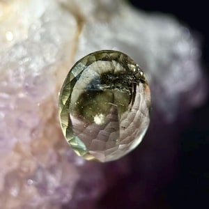 May include: A faceted, oval gemstone displaying a blend of green and clear hues. The gem features an intricate internal structure, set against a blurred, light-toned backdrop. The cut of the stone creates multiple reflective surfaces.