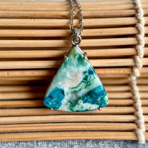 Puede incluir: Collar con cadena plateada y colgante triangular. El colgante es de piedra con tonos azules, verdes y blancos. La piedra está engastada en un marco plateado. El collar se exhibe sobre una superficie de bambú.