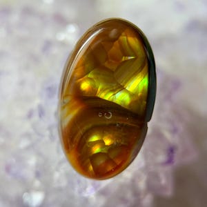 May include: A polished, oval-shaped gemstone with a vibrant display of colors. The gem features a mix of golden-brown, yellow, and green hues, with internal patterns that create a shimmering effect. The stone is set against a soft, light-colored background.