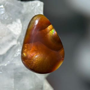 Op de afbeelding: Een gepolijste, druppelvormige edelsteen met warme amber- en oranje tinten. De steen heeft interne patronen en iriserende kleurflitsen, met een glad, glanzend oppervlak. De edelsteen is geplaatst tegen een vage, lichtgekleurde achtergrond.