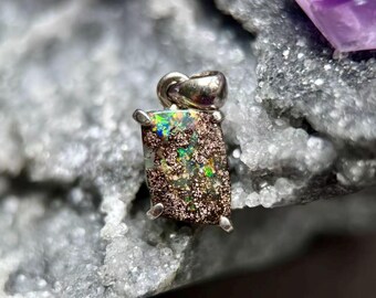 May include: A rectangular pendant featuring a dark, textured matrix stone with vibrant flashes of green, blue, and orange iridescent color. The stone is secured in a simple silver-toned metal setting with a small loop at the top for attachment.