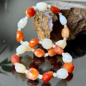 Op de afbeelding: Twee armbanden met facetgeslepen, ovale edelstenen in oranje, wit en perzik. De kralen zijn geregen met kleine, heldere kralen. De armbanden worden gepresenteerd op een reflecterend oppervlak.