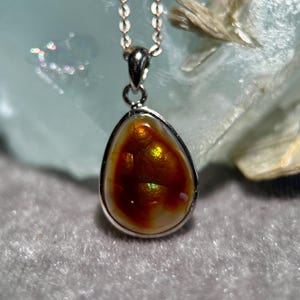 Op de afbeelding: Een druppelvormige hanger met een zilveren ketting. De hanger heeft een gepolijste, iriserende edelsteen in tinten oranje, bruin en goud, gezet in een zilveren bezel. De edelsteen reflecteert licht.
