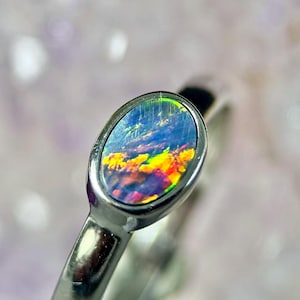 Könnte beinhalten: Ein silberner Ring mit einem ovalen Opal-Edelstein. Der Opal zeigt eine lebendige Farbpalette, darunter Blau, Gelb und Rot, was ein dynamisches und auffälliges Aussehen erzeugt. Der Ring hat ein poliertes Silberband.