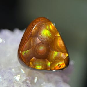 Op de afbeelding: Een gepolijste, driehoekige edelsteen met een vurige oranje en gele interne patroon. De edelsteen heeft een glad, reflecterend oppervlak en is geplaatst tegen een vage, lichtgekleurde achtergrond. Het ingewikkelde ontwerp van de steen wordt benadrukt door het spel van licht.