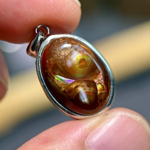 May include: An oval-shaped pendant featuring a polished fire agate gemstone set in a smooth silver-toned metal frame. The stone displays deep reddish-brown hues with iridescent flashes of gold, orange, and green light across its surface.