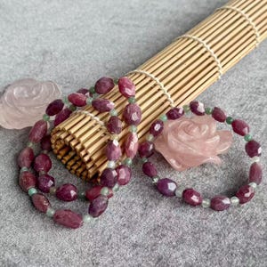 Könnte beinhalten: Nahaufnahme von zwei Halsketten und einem Armband aus facettierten, tiefroten Edelsteinen und kleinen grünen Perlen. Eine geschnitzte Rose aus rosa Quarz liegt neben dem Armband. Der Schmuck wird auf einer grauen Oberfläche mit einer Bambusmatte präsentiert.