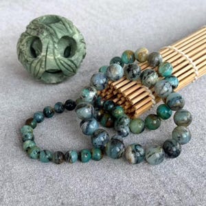 Pode incluir: Uma coleção de pulseiras de contas e uma esfera esculpida. As pulseiras apresentam contas redondas de vários tamanhos em tons de verde-azulado, verde e cinza. A esfera é uma pedra verde clara esculpida com um desenho floral. As contas são exibidas em uma esteira de bambu.