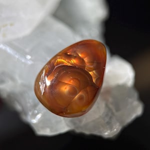 Op de afbeelding: Een gepolijste, druppelvormige edelsteen met wervelende patronen van oranje en bruin. De edelsteen is geplaatst tegen een achtergrond van wit, kristallijn materiaal, wat een opvallend contrast creëert. Het oppervlak van de steen reflecteert licht.