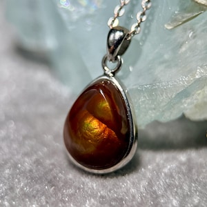 Op de afbeelding: Een zilveren hangerketting met een druppelvormige edelsteen, met levendige oranje en gouden tinten. De edelsteen is gezet in een zilveren rand en hangt aan een fijne zilveren ketting. De achtergrond heeft een zachte, vage textuur.