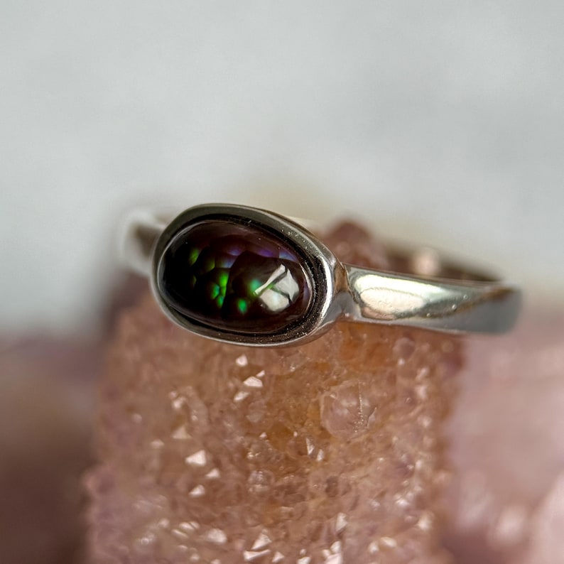 May include: A silver ring with an oval gemstone. The stone has a dark, iridescent quality, with dark brown, purple, and green colours. The ring is set on a light pink surface.