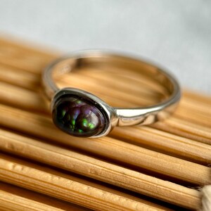 May include: A silver ring with an oval gemstone displaying a mosaic of colours: green, purple, and black. The ring has a polished silver band. The gemstone is the central feature, exhibiting a unique, iridescent pattern.