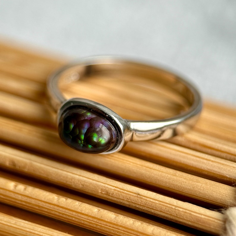 May include: A silver ring with an oval gemstone displaying a mosaic of colours: green, purple, and black. The ring has a polished silver band. The gemstone is the central feature, exhibiting a unique, iridescent pattern.