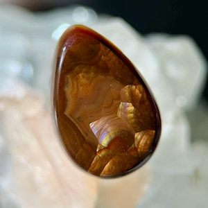 Op de afbeelding: Een gepolijste, druppelvormige edelsteen met een rijk, aards kleurenpalet. De steen heeft wervelende patronen van bruin, oranje en goud, met gebieden van iriserende glans. Het oppervlak van de edelsteen is glad en reflecterend.