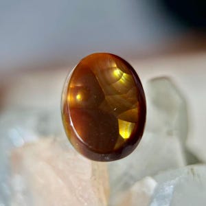 Op de afbeelding: Een gepolijste, ovaalvormige edelsteen met een warme bruine kleur en interne goudgele highlights. De edelsteen heeft een glad, reflecterend oppervlak, met natuurlijke patronen. De steen is geplaatst tegen een zachte, vage achtergrond.