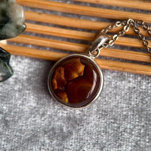 Op de afbeelding: Een ronde hangerketting met een zilverkleurige ketting. De hanger heeft een cirkelvormige, gepolijste edelsteen met bruine en gouden tinten. De edelsteen is gezet in een zilveren rand. De achtergrond omvat een houten oppervlak en een grijze, getextureerde stof.