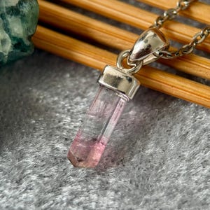May include: A pendant featuring a translucent, elongated, prismatic crystal with a gradient color transition from clear at the top to a soft pink at the base. The crystal is secured by a polished silver-toned metal cap and attached to a silver-toned chain.