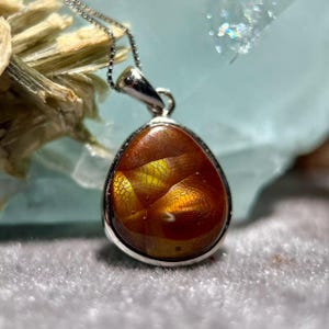Op de afbeelding: Een zilveren hangerketting met een druppelvormige edelsteen. De edelsteen heeft een amberkleur met interne patronen van geel en oranje. De hanger is gezet in een zilveren rand en hangt aan een fijne zilveren ketting.