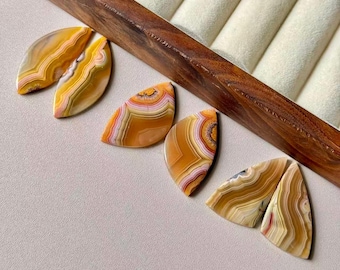 May include: Six polished agate gemstones in shades of yellow, orange, and white, with unique banded patterns. The gemstones are displayed on a light gray surface near a wooden jewelry tray.