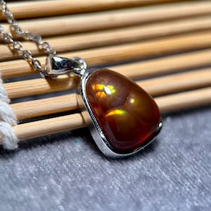Op de afbeelding: Een zilveren hangerketting met een druppelvormige, gepolijste edelsteen. De edelsteen is een rijk, warm bruin met interne flitsen van geel en oranje. De hanger is gezet in een zilveren rand en hangt aan een fijne zilveren ketting.