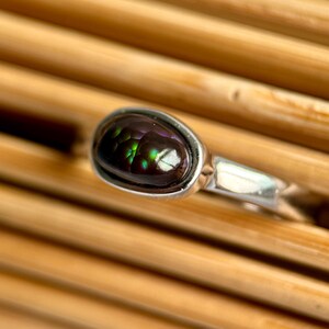 May include: A silver ring with a dark gemstone, displaying purple, green, and brown colours. The oval stone is set in a simple silver band. The ring is shown on a wooden surface.